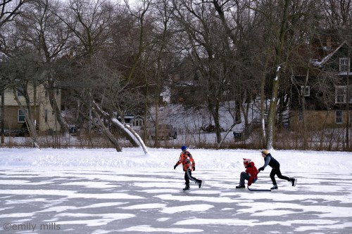 Three children skate on a frozen lake, surrounded by snow-covered trees and houses in the background.