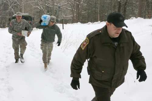 Three individuals walk through snow, one carrying water bottles, while another wears a uniform and the third is in a police jacket.