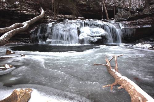 A frozen waterfall surrounded by icy rocks and a snowy landscape, with a partially frozen pool in the foreground.