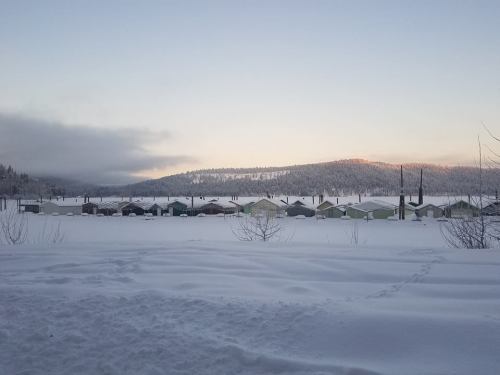 Snow-covered landscape with colorful cabins in the distance, surrounded by mountains and a clear sky at dawn.