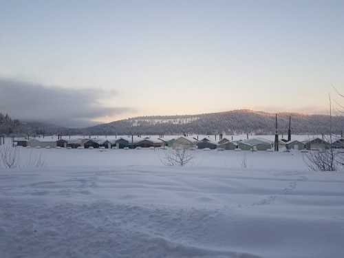 Snow-covered landscape with colorful cabins in the distance, surrounded by mountains and a clear sky at dawn.