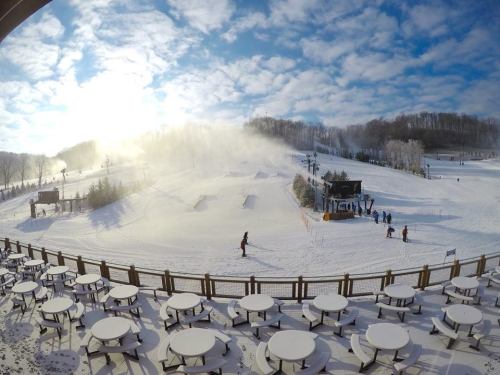 A snowy ski resort scene with skiers, snow-making machines, and empty tables on a deck under a cloudy sky.