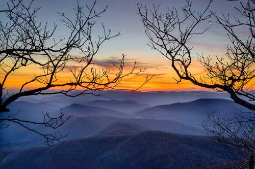 A serene sunset over misty mountains, framed by bare tree branches against a colorful sky.