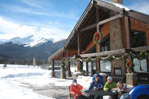 A cozy mountain lodge surrounded by snow, with people enjoying drinks outside and festive decorations.