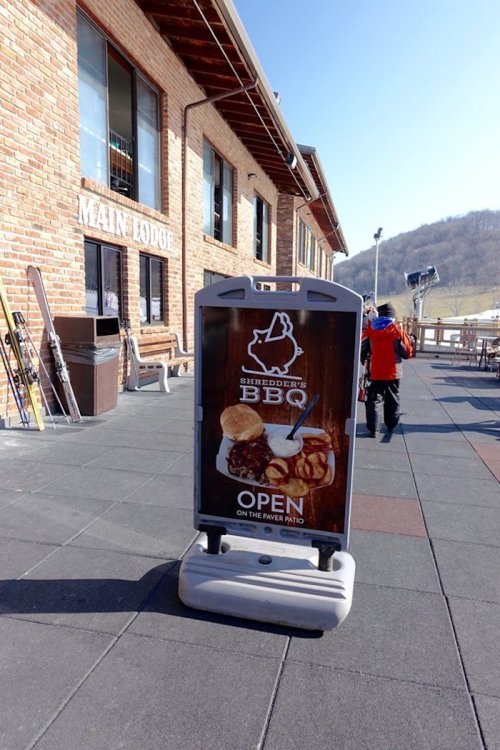A sign for Shedders BBQ outside the Maine Lodge, indicating it's open on the river patio, with a snowy landscape in the background.
