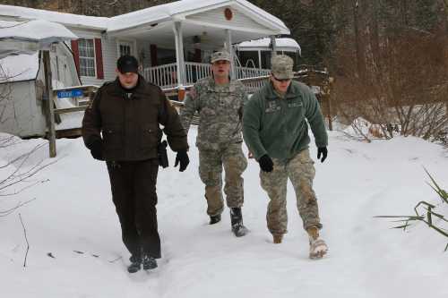 Three men in military and law enforcement attire walking through snow near a house.