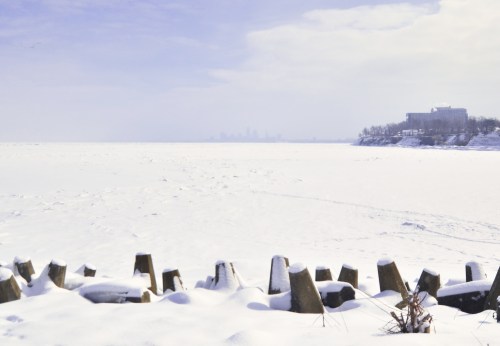 A snowy landscape with frozen water, wooden posts in the foreground, and a distant city skyline under a cloudy sky.