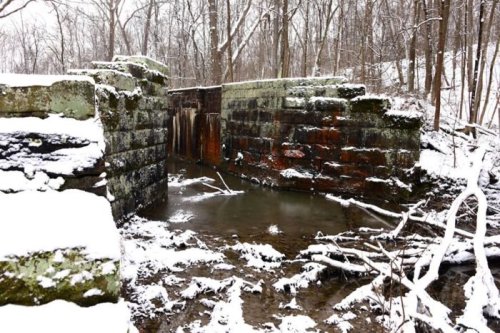 Snow-covered stone ruins by a frozen waterway, surrounded by bare trees in a winter landscape.