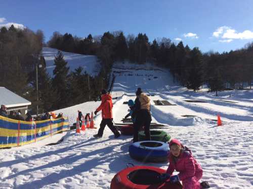 Children and adults enjoy snow tubing on a sunny winter day, with snowy hills and trees in the background.