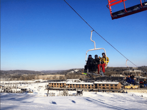 Skiers on a chairlift with a snowy landscape and a lodge in the background under a clear blue sky.