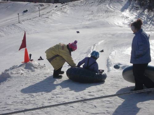 A snowy hill with people tubing; one person helps another onto a tube while a third person watches nearby.