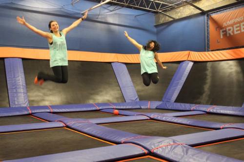 Two women jump joyfully on a trampoline, wearing matching teal tank tops, in a vibrant indoor trampoline park.