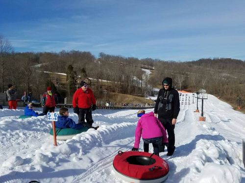A snowy tubing hill with people preparing to slide down, surrounded by trees and a clear blue sky.