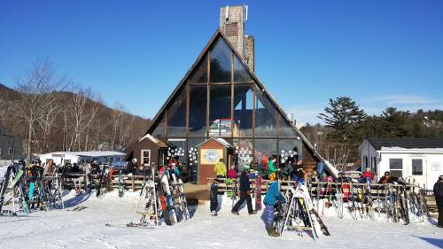A ski lodge with an A-frame design, surrounded by snow and skiers, with equipment lined up outside on a sunny day.