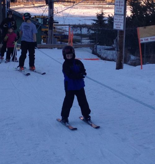 A child in ski gear stands on a snowy slope, ready to ski, with others in the background waiting in line.