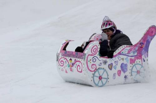 A child and an adult ride a colorful, decorated sled down a snowy slope, both wearing helmets and smiling.