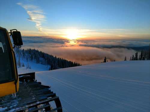 A snow-covered landscape at sunset, with clouds below and a snow groomer in the foreground.