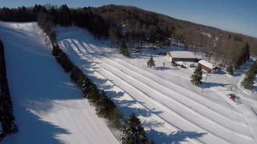 Aerial view of a snowy landscape with groomed ski trails and a small building surrounded by trees.