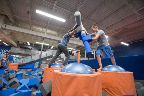 Two kids play on a foam pit, using large foam poles to balance and compete in a fun, active environment.