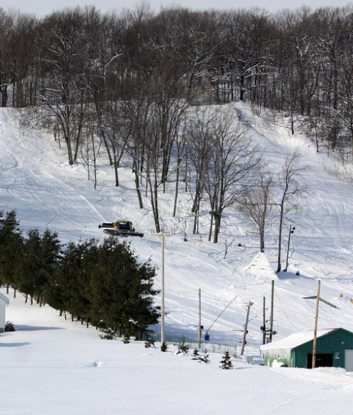 A snow-covered landscape with trees, a ski slope, and a snow grooming machine in the distance.