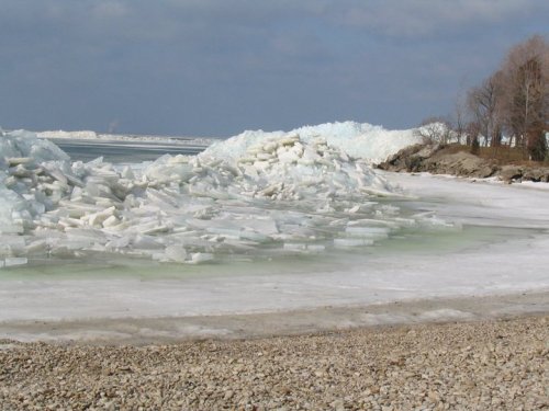 A frozen shoreline with large ice formations and a cloudy sky, featuring a mix of ice and gravel along the water's edge.