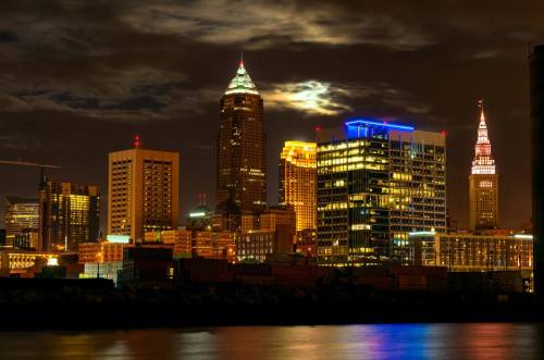 Cleveland skyline at night, featuring illuminated skyscrapers and a cloudy sky reflected in the water.