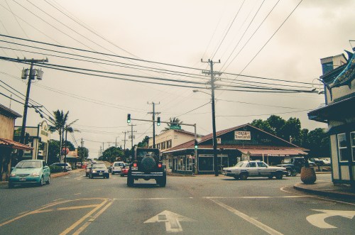 A street scene with cars, traffic lights, and shops under a cloudy sky. Power lines crisscross above.