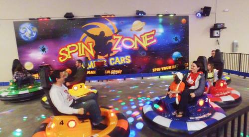A colorful indoor amusement area with people enjoying bumper cars under vibrant lights and a "Spin Zone" backdrop.