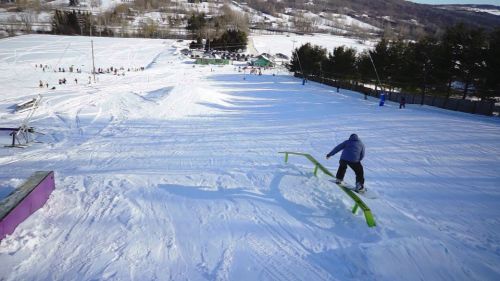 A snowboarder performs a trick on a green rail in a snowy landscape with hills and people in the background.