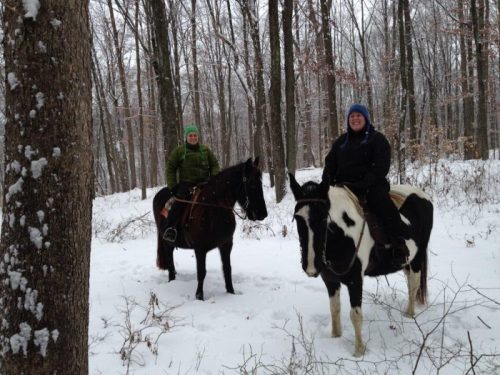 Two people on horseback in a snowy forest, surrounded by trees, wearing winter clothing and smiling.