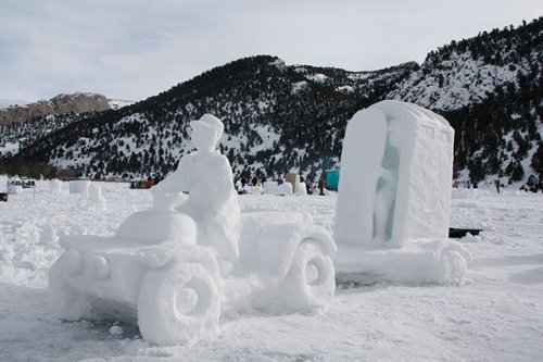 A snow sculpture of a person riding an ATV, pulling a snow structure resembling a portable restroom. Snowy mountains in the background.
