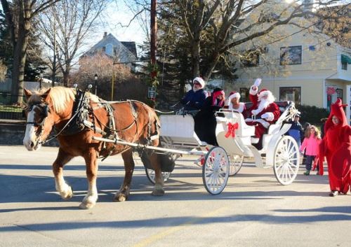 A festive horse-drawn carriage with Santa and his helpers waves to a crowd during a holiday parade.