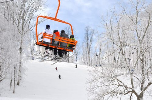 A ski lift carries passengers over a snowy landscape with skiers on the slope below and trees in the background.