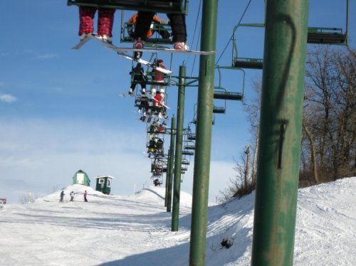 Skiers on a chairlift ascend a snowy slope under a clear blue sky, with a small building in the background.