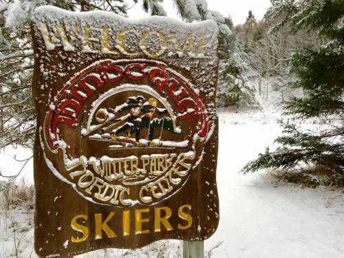 A snowy sign reading "Welcome Skiers" at the Minnesota Winter Park Nordic Center, surrounded by trees.