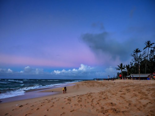 A serene beach at dusk with gentle waves, palm trees, and a person walking along the sandy shore.