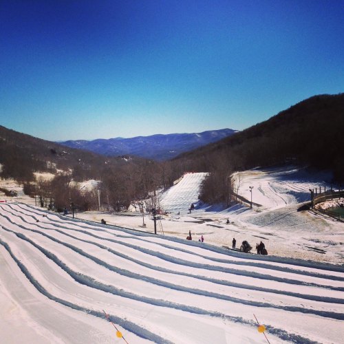 A snowy landscape with multiple sledding lanes, surrounded by hills and a clear blue sky. People enjoy winter activities.