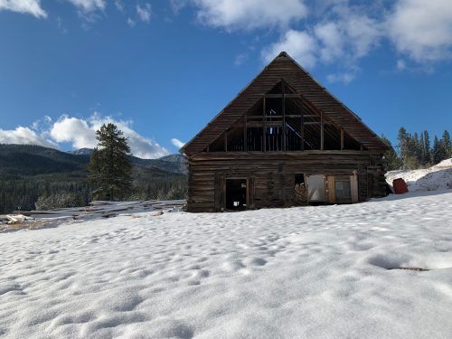 A rustic wooden barn in a snowy landscape, surrounded by mountains and a clear blue sky.