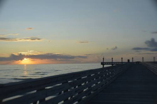 A wooden pier extends into the ocean at sunset, with soft clouds and warm colors in the sky.