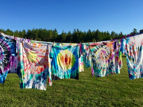 Colorful tie-dye shirts hanging on a clothesline against a clear blue sky and green trees in the background.
