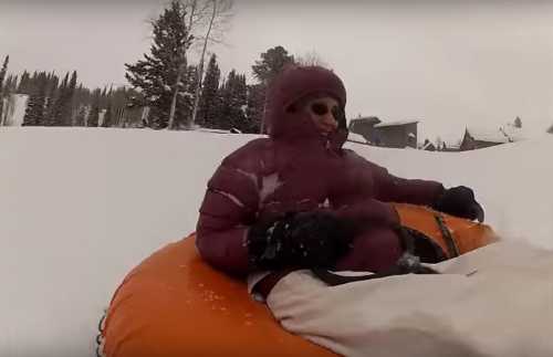 A person in a purple winter coat sits in an orange snow tube, surrounded by a snowy landscape and trees.