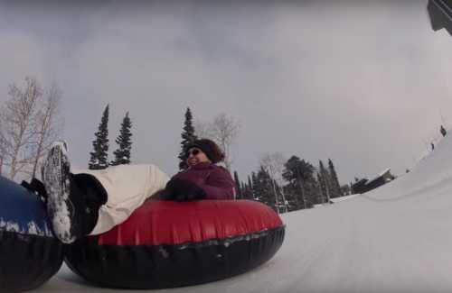 A person in winter clothing enjoys tubing down a snowy slope, surrounded by trees and a cloudy sky.