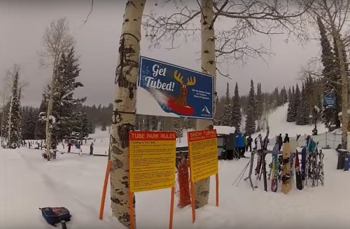 A snowy landscape with a "Get Tubed!" sign, surrounded by trees and people enjoying winter sports.
