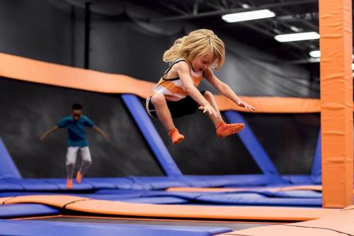 A child jumps joyfully on a trampoline in a vibrant indoor trampoline park. Another child is seen in the background.