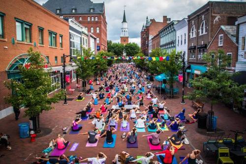 A large group of people practicing yoga outdoors on colorful mats in a bustling town square, with a church steeple in the background.