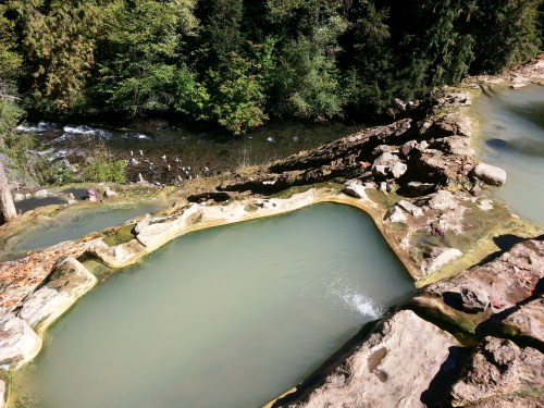 Natural hot springs with terraced pools, surrounded by lush greenery and a flowing river in the background.