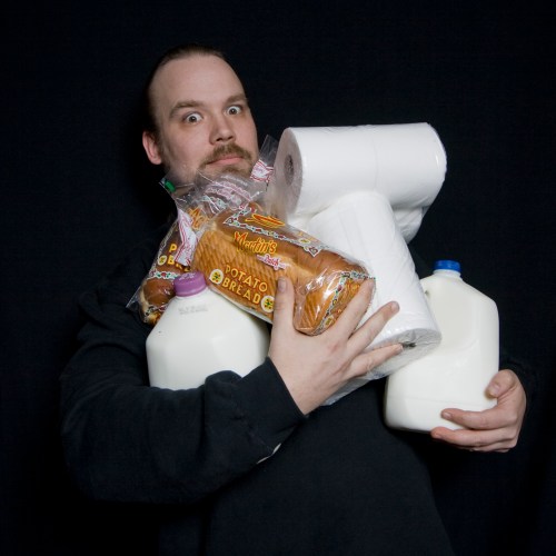 A person holds multiple grocery items, including milk jugs, bread, and toilet paper, with a surprised expression.
