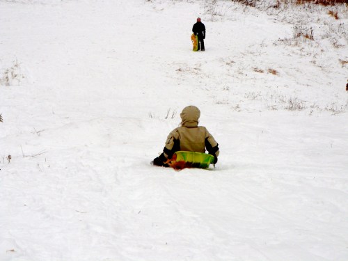 A child sleds down a snowy hill while another person stands at the top, ready to slide down.