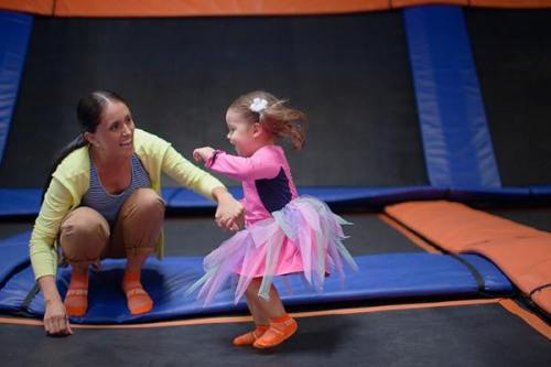 A woman assists a young girl in a pink tutu as they jump on a trampoline, both smiling and enjoying their time together.