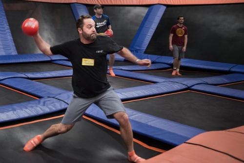 A man throws a dodgeball on a trampoline court, with two others in the background preparing to play.